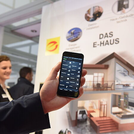 Es ist eine Hand zu sehen, die ein Handy mit einer App in die Kamera hält. Im Hintergrund ist eine Frau zu erkennen und eine Informationswand zu sehen auf der die Beschriftung Das E-Haus steht und weitere Informationen.