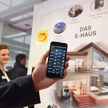 Es ist eine Hand zu sehen, die ein Handy mit einer App in die Kamera hält. Im Hintergrund ist eine Frau zu erkennen und eine Informationswand zu sehen auf der die Beschriftung Das E-Haus steht und weitere Informationen.
