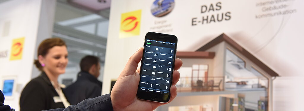 Es ist eine Hand zu sehen, die ein Handy mit einer App in die Kamera hält. Im Hintergrund ist eine Frau zu erkennen und eine Informationswand zu sehen auf der die Beschriftung Das E-Haus steht und weitere Informationen.