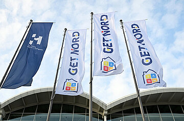 Fahnen mit dem Logo der Messe GET NORD vor dem Eingangsbereich de Hamburg Messe unter blauem Himmel.