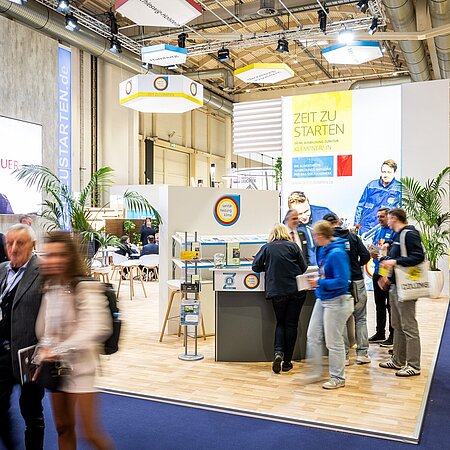  Messestand zum Thema „Sanitär, Heizung, Klima“ mit großem Bildschirm und dem Hashtag „#WICHTIGESTUER“. Ein Schild zeigt „Zeit zu starten“, Personen sind im Gespräch oder laufen vorbei.