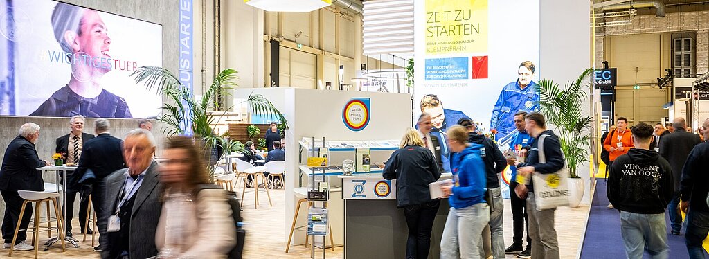  Messestand zum Thema „Sanitär, Heizung, Klima“ mit großem Bildschirm und dem Hashtag „#WICHTIGESTUER“. Ein Schild zeigt „Zeit zu starten“, Personen sind im Gespräch oder laufen vorbei.