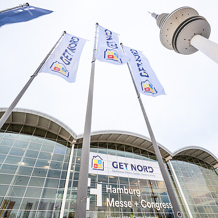 Blick von unten auf mehrere Fahnen mit der Aufschrift „GET NORD“, die vor dem Gebäude der Hamburg Messe + Congress im Wind wehen; im Hintergrund sind die Glasfassade, der Eingangsbereich mit Beschriftung sowie ein hoher Fernsehturm und eine Straßenleuchte zu sehen.