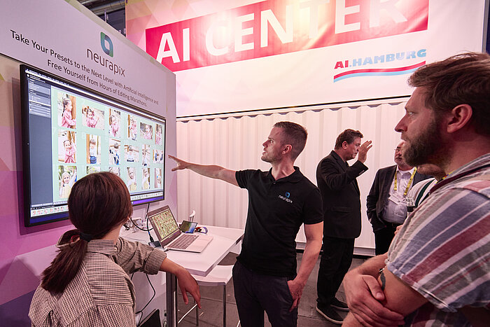 PHOTOPIA 2023 – Messeimpressionen AI CENTER Ein Mann demonstriert auf einem Bildschirm die Software „Neurapix“, die mithilfe von künstlicher Intelligenz Fotobearbeitungsprozesse optimiert, während mehrere Personen ihm aufmerksam folgen.