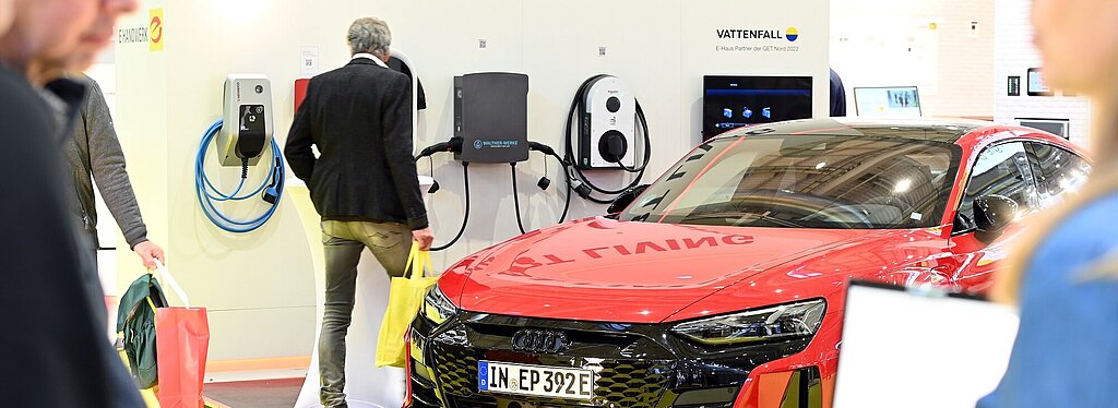 Es ist ein rotes E-Auto bei einer Ladestation von Vattenfall zu sehen. An einer Wand steht Smart Living und Personen laufen im Innenraum umher. 