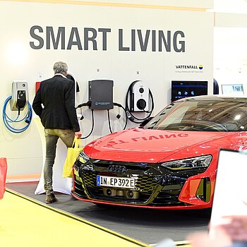 Es ist ein rotes E-Auto bei einer Ladestation von Vattenfall zu sehen. An einer Wand steht Smart Living und Personen laufen im Innenraum umher. 
