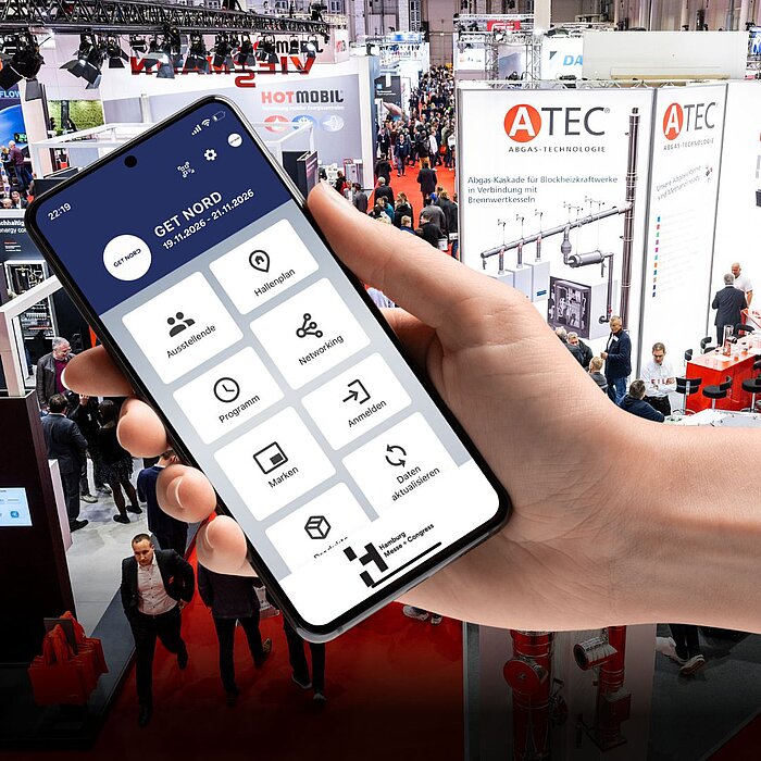 Eine Hand hält ein Smartphone. Auf dem Bildschirm ist der Homescreen der GET NORD App zu sehen mit den Bereichen "Ausstellende", "Hallenplan", "Programm", "Networking", "Marken" und "Anmelden".. Im Hintergrund sieht man eine Szene aus der laufenden Messebetrieb der GET NORD.