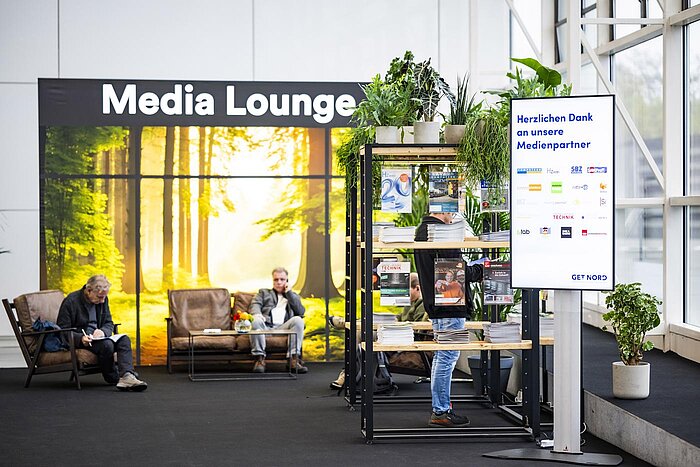 Blick von Eingang Mitte auf die Media Lounge. Mehrere gemütliche Sessel stehen vor einer Wand mit Naturdruck und dem Schriftzug Media Lounge. Daneben stehen einige Regale mit Zeitschriften der Medienpartner.