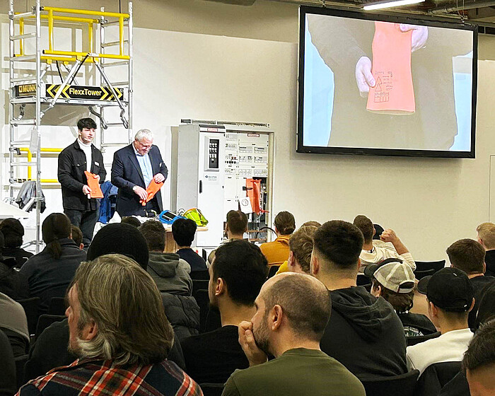 Zwei Sprecher halten eine Präsentation vor einem Publikum im Rahmen eines Arbeitssicherheitsseminars, mit Sicherheitshandschuhen in der Hand und einem Bildschirm im Hintergrund.