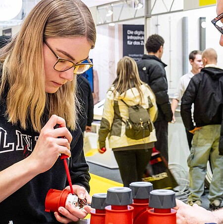 Eine junge Frau auf der linken Bildseite öffnet konzentriert mit einem Schraubenschlüssel ein kleines Gerät. Ein Mann auf der rechten Seite unterstützt sie dabei.