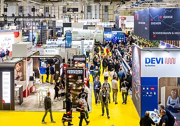 Innenaufnahme einer Messehalle mit zahlreichen Ausstellern und Besuchern. Die Halle ist hell beleuchtet, mit gelbem Boden. Zu sehen sind zahlreiche Stände.