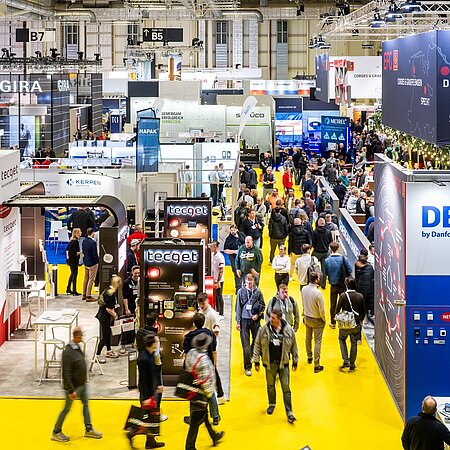 Innenaufnahme einer Messehalle mit zahlreichen Ausstellern und Besuchern. Die Halle ist hell beleuchtet, mit gelbem Boden. Zu sehen sind zahlreiche Stände.